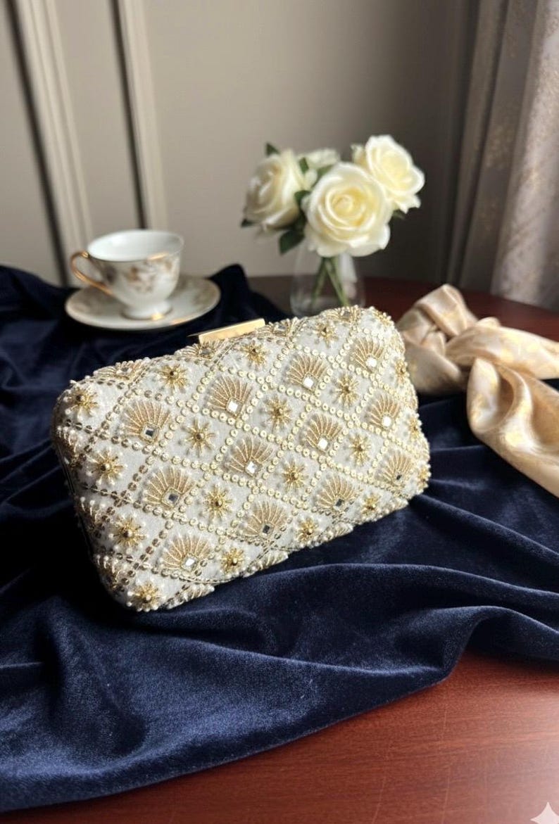 May include: Ivory rectangular clutch bag with gold and pearl detailing. The beaded design has a geometric pattern with fan and starburst shapes. A gold clasp secures the top. The bag sits on a dark blue velvet surface.