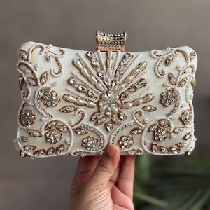 May include: Ivory rectangular clutch with intricate beadwork and embellishments. The clutch features a central sunburst design with clear crystals and pearls, surrounded by floral patterns and gold accents. A gold clasp secures the top.