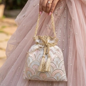Puede incluir: Un bolso con cordón de color marfil con un asa de cuentas doradas y borlas. El bolso presenta un patrón bordado en tonos rosa, dorado y plateado. Se sostiene sobre un fondo de tela rosa suave.