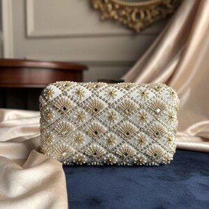 May include: White rectangular clutch bag with detailed gold and pearl beadwork. The design includes a diamond grid pattern with floral and fan-shaped elements. A small, rectangular clasp secures the top.