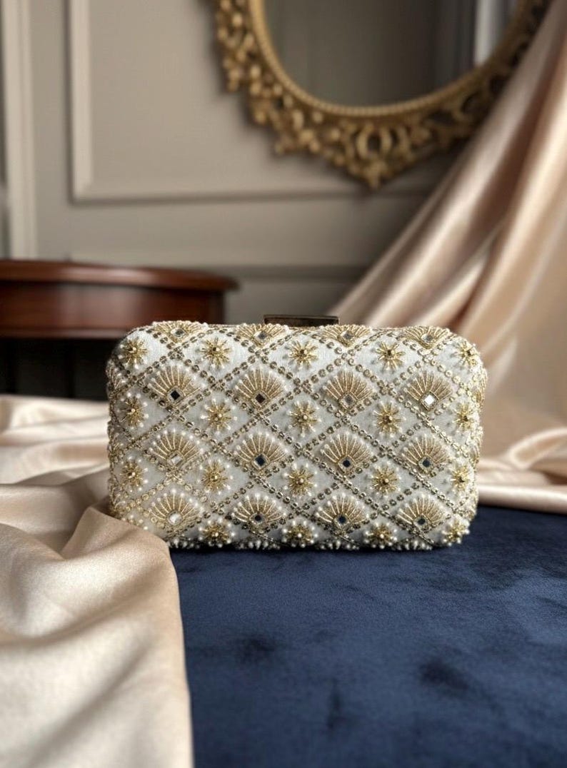 May include: White rectangular clutch bag with detailed gold and pearl beadwork. The design includes a diamond grid pattern with floral and fan-shaped elements. A small, rectangular clasp secures the top.