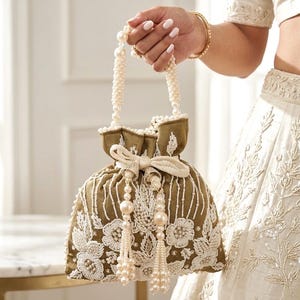 May include: An olive green drawstring bag with white floral embroidery and pearl accents. The bag features a pearl beaded handle and tassel details. The bag is held by a person wearing a gold bracelet.