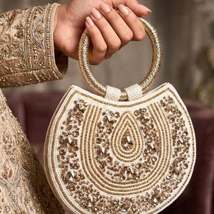 Könnte beinhalten: Eine weiße und goldfarbene, mit Perlen besetzte Handtasche mit einem kreisförmigen goldenen Griff. Die Tasche weist aufwendige goldfarbene Perlenarbeiten und Verzierungen auf. Der Griff ist ein dicker, goldfarbener Ring. Die Tasche wird von einer Person gehalten, die ein goldfarbenes, besticktes Kleidungsstück trägt.