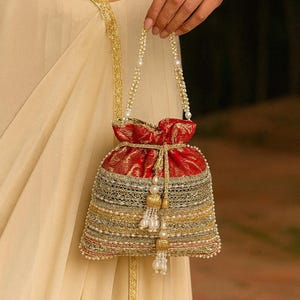 Puede incluir: Un bolso rojo y dorado con cordón y adornos de cuentas y perlas. El bolso presenta un asa y borlas con cuentas de oro y perlas. El bolso lo sostiene una persona que lleva un atuendo color crema con ribetes dorados.