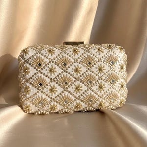May include: Ivory rectangular clutch bag with detailed gold bead and mirror embellishments. The bag has a geometric pattern of diamonds, fans, and starbursts. The clasp is gold-toned, set against a satin fabric background.