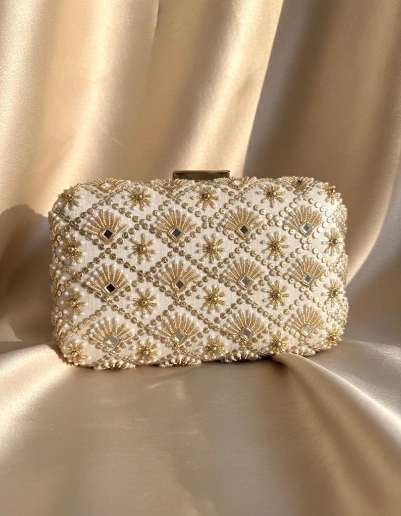 May include: Ivory rectangular clutch bag with detailed gold bead and mirror embellishments. The bag has a geometric pattern of diamonds, fans, and starbursts. The clasp is gold-toned, set against a satin fabric background.