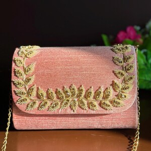 May include: A blush pink clutch with a gold chain strap. The flap is adorned with gold-colored leaf-shaped embellishments. The bag is made of a textured fabric and has a rectangular shape.