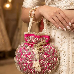 May include: A hot pink, round, embroidered purse with a pearl handle and tassel. The purse is adorned with silver and white floral embroidery and a gold trim. The purse is held by a person wearing a white outfit and pearl bracelets.