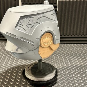 K&ouml;nnte beinhalten: Ein grau-beiger Helm mit aufwendigen Schnitzereien, darunter ein baumartiges Design an der Seite. Der Helm ist auf einem St&auml;nder montiert, der sein Design und seine Handwerkskunst zeigt. Der Sockel ist schwarz und die Oberfl&auml;che metallisch.