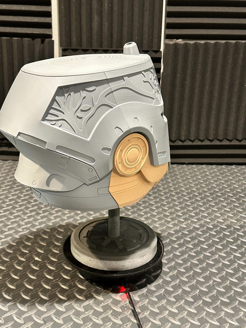 K&ouml;nnte beinhalten: Ein grau-beiger Helm mit aufwendigen Schnitzereien, darunter ein baumartiges Design an der Seite. Der Helm ist auf einem St&auml;nder montiert, der sein Design und seine Handwerkskunst zeigt. Der Sockel ist schwarz und die Oberfl&auml;che metallisch.