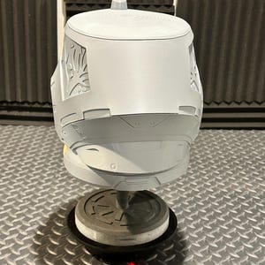 K&ouml;nnte beinhalten: Ein grauer, 3D-gedruckter Helm mit aufwendigen Designs an den Seiten. Der Helm ist auf einem St&auml;nder mit kreisf&ouml;rmiger Basis vor einem strukturierten Hintergrund montiert. Das Helmdesign weist eine obere Finne und detaillierte Oberfl&auml;chenelemente auf.