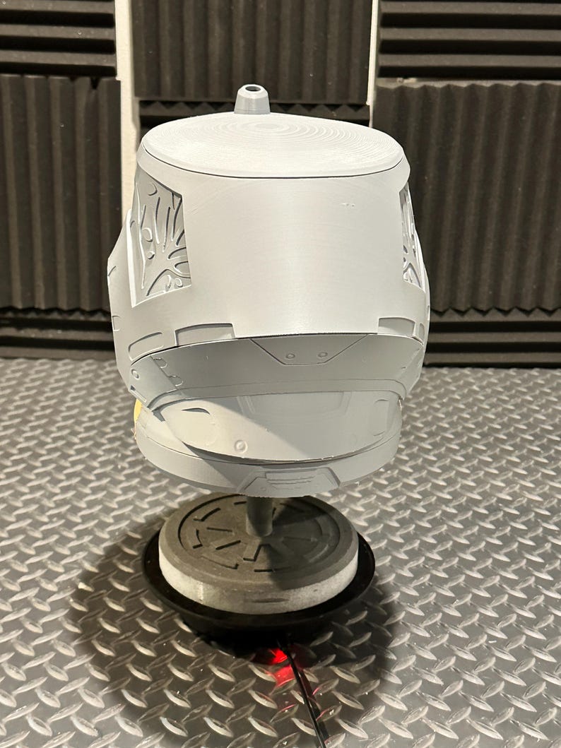K&ouml;nnte beinhalten: Ein grauer, 3D-gedruckter Helm mit aufwendigen Designs an den Seiten. Der Helm ist auf einem St&auml;nder mit kreisf&ouml;rmiger Basis vor einem strukturierten Hintergrund montiert. Das Helmdesign weist eine obere Finne und detaillierte Oberfl&auml;chenelemente auf.