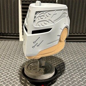 K&ouml;nnte beinhalten: Ein grau-beiger Helm mit aufwendigen Schnitzereien, darunter ein stilisiertes Bl&auml;ttermuster und drei kleine parallele Linien. Der Helm wird auf einem St&auml;nder pr&auml;sentiert, der sein Design hervorhebt. Die Gesamt&auml;sthetik deutet auf ein futuristisches oder Fantasy-Thema hin.