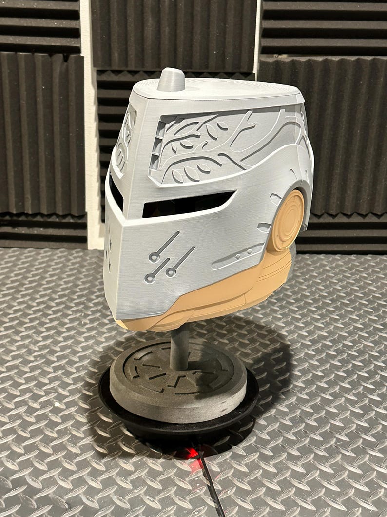 K&ouml;nnte beinhalten: Ein grau-beiger Helm mit aufwendigen Schnitzereien, darunter ein stilisiertes Bl&auml;ttermuster und drei kleine parallele Linien. Der Helm wird auf einem St&auml;nder pr&auml;sentiert, der sein Design hervorhebt. Die Gesamt&auml;sthetik deutet auf ein futuristisches oder Fantasy-Thema hin.
