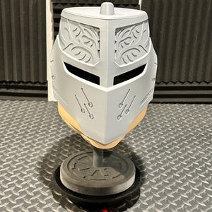 K&ouml;nnte beinhalten: Ein hellgrauer, 3D-gedruckter Helm mit dekorativen Bl&auml;ttermustern auf der Oberseite und den Seiten. Der Helm hat Augenschlitze und ist auf einem grauen St&auml;nder mit kreisf&ouml;rmiger Basis montiert. Der Helm wird auf einer Riffelblechoberfl&auml;che ausgestellt.