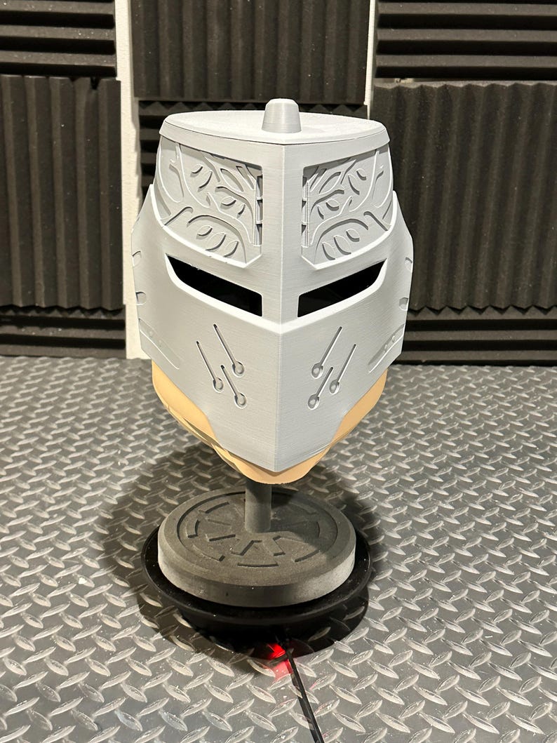K&ouml;nnte beinhalten: Ein hellgrauer, 3D-gedruckter Helm mit dekorativen Bl&auml;ttermustern auf der Oberseite und den Seiten. Der Helm hat Augenschlitze und ist auf einem grauen St&auml;nder mit kreisf&ouml;rmiger Basis montiert. Der Helm wird auf einer Riffelblechoberfl&auml;che ausgestellt.