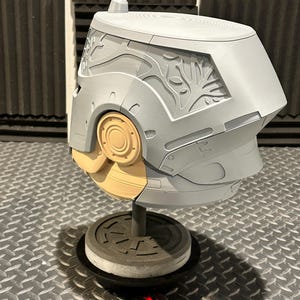 K&ouml;nnte beinhalten: Ein grauer und beigefarbener Helm mit aufwendigen Schnitzereien, darunter ein baumartiges Design an der Seite. Der Helm ist auf einem St&auml;nder mit kreisf&ouml;rmiger Basis montiert. Das Design weist glatte und strukturierte Oberfl&auml;chen auf.