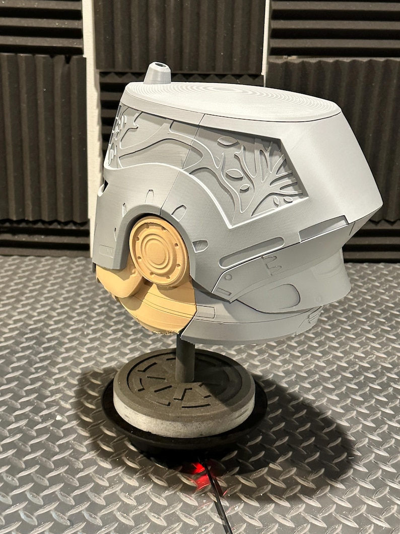 K&ouml;nnte beinhalten: Ein grauer und beigefarbener Helm mit aufwendigen Schnitzereien, darunter ein baumartiges Design an der Seite. Der Helm ist auf einem St&auml;nder mit kreisf&ouml;rmiger Basis montiert. Das Design weist glatte und strukturierte Oberfl&auml;chen auf.