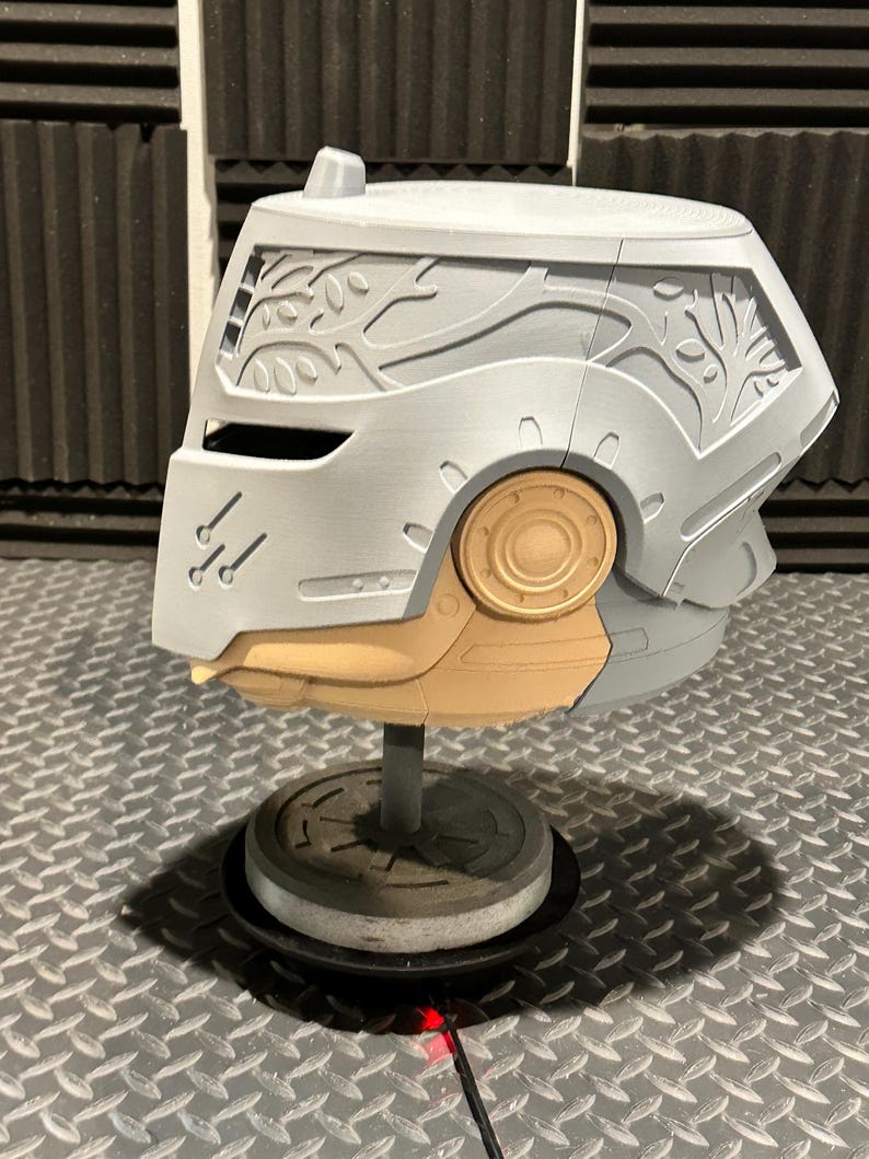 K&ouml;nnte beinhalten: Ein grauer und beigefarbener Helm mit aufwendigen Baumzweigmustern auf den Seitenteilen. Der Helm hat ein kreisf&ouml;rmiges, goldfarbenes Detail und ist auf einem grauen St&auml;nder montiert. Der Sockel ist kreisf&ouml;rmig und steht auf einer metallischen Oberfl&auml;che mit Rautenmuster.