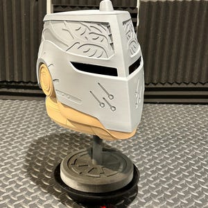 K&ouml;nnte beinhalten: Ein hellgrauer Helm mit Visier und dekorativen Bl&auml;ttermustern. Der Helm wird auf einem St&auml;nder mit kreisf&ouml;rmiger Basis ausgestellt. Der Helm hat einen hellbraunen unteren Teil und eine kleine zylindrische Oberseite.