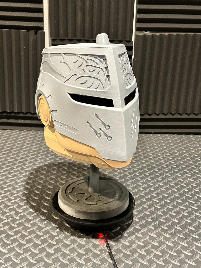 K&ouml;nnte beinhalten: Ein hellgrauer Helm mit Visier und dekorativen Bl&auml;ttermustern. Der Helm wird auf einem St&auml;nder mit kreisf&ouml;rmiger Basis ausgestellt. Der Helm hat einen hellbraunen unteren Teil und eine kleine zylindrische Oberseite.