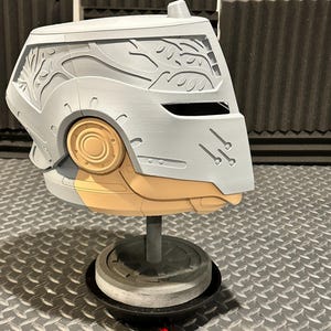 K&ouml;nnte beinhalten: Ein grauer und beigefarbener Helm mit aufwendigen Schnitzereien, darunter ein Astmuster an der Seite. Der Helm ist auf einem grauen St&auml;nder montiert, der sein futuristisches Design hervorhebt. Der Helm hat ein kreisf&ouml;rmiges, goldfarbenes Element an der Seite.