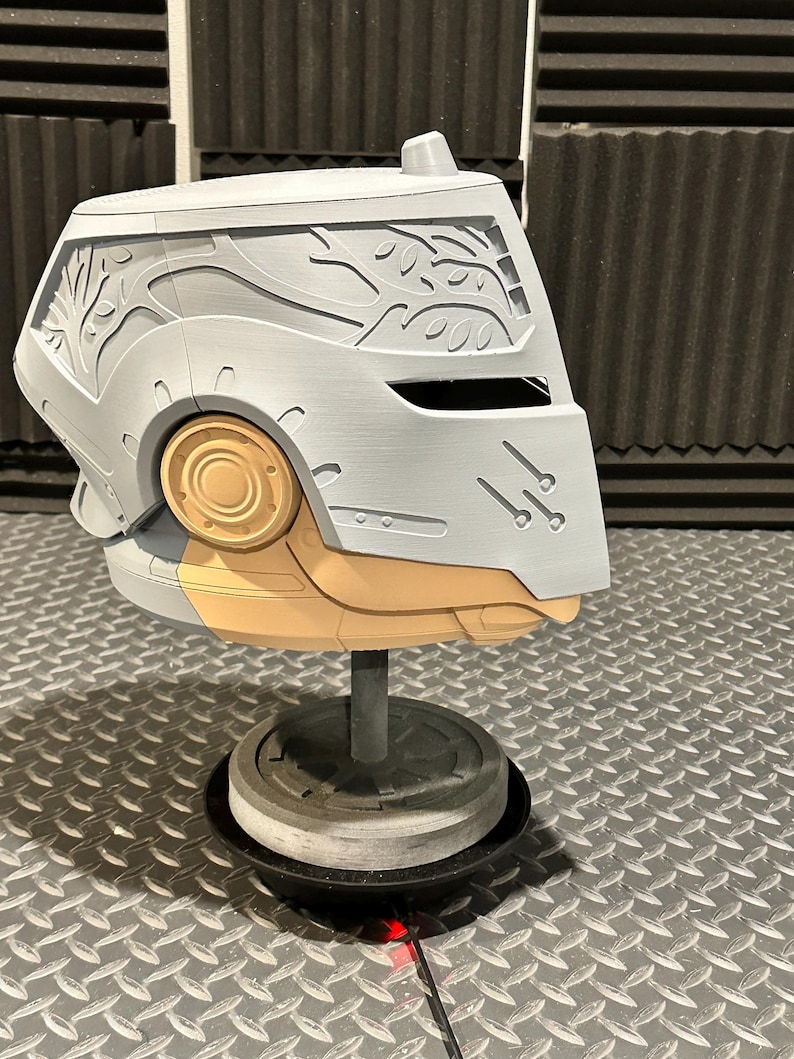 K&ouml;nnte beinhalten: Ein grauer und beigefarbener Helm mit aufwendigen Schnitzereien, darunter ein Astmuster an der Seite. Der Helm ist auf einem grauen St&auml;nder montiert, der sein futuristisches Design hervorhebt. Der Helm hat ein kreisf&ouml;rmiges, goldfarbenes Element an der Seite.