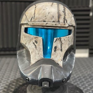 Puede incluir: Un casco negro y gris desgastado con una visera azul. El casco tiene un aspecto desgastado y deteriorado, con arañazos y rozaduras visibles.