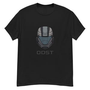 Può includere: T-shirt nera con grafica di un casco stilizzato con accenti grigi e blu. La parola "ODST" è stampata in grigio sotto il disegno del casco. La maglietta è realizzata in un materiale morbido e confortevole.