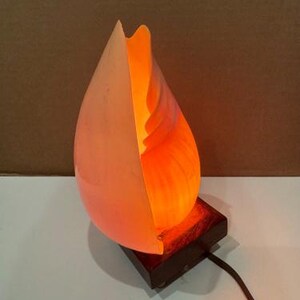 Puede incluir: Lámpara de vidrio artístico con un diseño único. La lámpara tiene forma de lágrima, con tonos blancos y naranjas. Se asienta sobre una base de madera marrón oscuro y tiene un cable de alimentación negro. La lámpara está encendida.