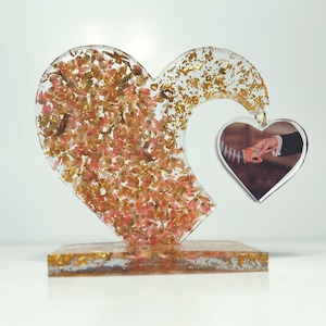 May include: A clear resin heart-shaped sculpture with gold glitter and pink flowers embedded inside. The sculpture has a smaller heart-shaped cutout that holds a photo of a couple holding hands.