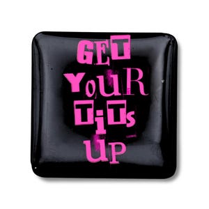 May include: A square, black button with the phrase "GET YOUR TITS UP" in bright pink, bold letters. The text is centered and fills the button's surface. The button has a glossy finish and rounded corners.