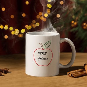 Può includere: Una tazza da caffè in ceramica bianca con un design a forma di mela rossa. La mela ha una foglia verde e il testo "MRS. Johnson" all'interno.