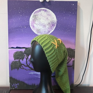 May include: A long, green, crocheted hat with a ribbed brim and a pointed, drooping tail. The hat features two yellow, cross-stitched accents on the side. It is displayed on a black, featureless mannequin head, showcasing the texture of the yarn.