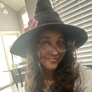 May include: A dark grey crocheted witch hat with a wide brim and a pointed crown. The hat is decorated with a pink and green crocheted flower. This handmade hat is ideal for Halloween or fancy dress.