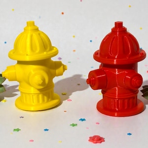May include: A yellow and a red fire hydrant, side by side, on a white surface. The hydrants have a classic design with a rounded top and multiple nozzles. Green succulents and colorful confetti surround the hydrants.