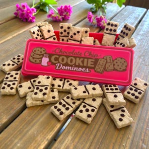 May include: A pink rectangular box with the words "Chocolate Chip COOKIE Dominoes" contains cookie-shaped dominoes. The dominoes are beige with dark brown dots. The box is on a wooden surface with pink flowers.