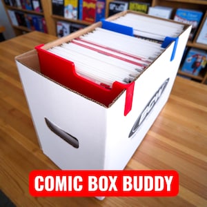 Puede incluir: Caja de almacenamiento de cómics de cartón blanco con divisores rojos y azules. La caja está llena de cómics y tiene el texto "COMIC BOX BUDDY" en una pancarta roja en la parte inferior. La caja tiene un recorte de asa en el lateral.