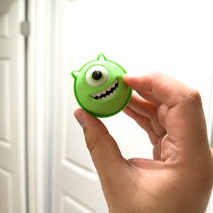 Puede incluir: Un personaje de dibujos animados verde y circular con un solo ojo, una gran sonrisa y pequeños cuernos. El personaje se sostiene en una mano. El diseño es probablemente de una película o serie animada.