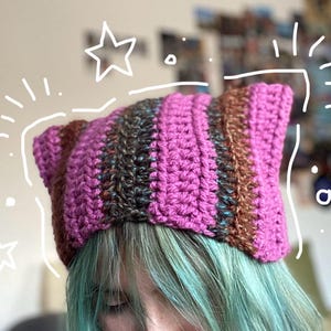 May include: A handmade crochet cat ear hat in shades of pink, brown, and dark grey. The hat features a striped pattern and is designed with pointed cat ears. The hat is a unique accessory.