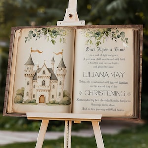 May include: A wooden easel displays a sign with a fairytale castle illustration and text. The sign reads "Once Upon a Time" and "LILIANA MAY CHRISTENING." The sign is 36 inches by 24 inches. The background is a blurred outdoor scene.