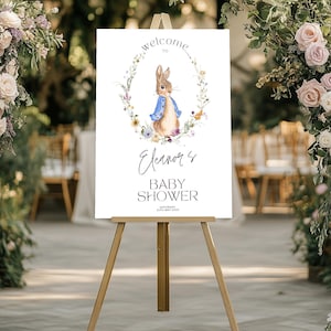 Può includere: Un cartello di benvenuto per la Baby Shower di Eleanor con sfondo bianco e una ghirlanda di fiori con un coniglio al centro. Il coniglio indossa una giacca blu ed è circondato da fiori. Il testo "Benvenuti alla Baby Shower di Eleanor" è scritto in corsivo nero.