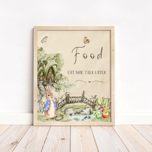 May include: A watercolour illustration of a rabbit wearing a blue jacket standing in front of a small wooden bridge. The illustration is framed in a light wood frame and has the text "Food Eat Now, Talk Later" on a beige background.