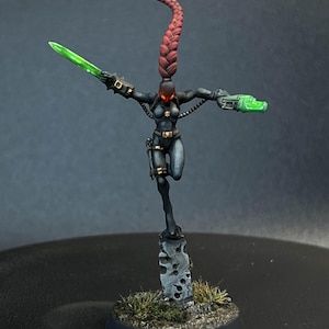 May include: Miniature figure of a female warrior in a black bodysuit, holding a green energy sword and a green energy pistol. The figure has a long, red braided ponytail and is standing on a stone base with grass.