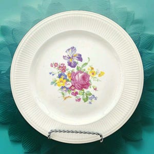 Puede incluir: Plato de cerámica blanca con un diseño floral que presenta una rosa rosa, un iris púrpura y otras flores coloridas. El plato tiene un borde acanalado y se exhibe sobre un soporte de metal plateado.