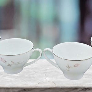 Könnte beinhalten: Vier weiße Teetassen mit zartem Blumendesign und silberner Verzierung. Jede Tasse hat einen Henkel und einen leicht abgewinkelten Rand. Die Tassen sind auf einer marmorähnlichen Oberfläche angeordnet.