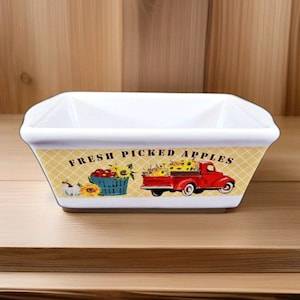 May include: White rectangular ceramic dish with a yellow checkered border. The border features a red truck, a basket of sunflowers, and the text "FRESH PICKED APPLES". The dish is on a wooden surface.