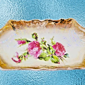 Puede incluir: Una fuente rectangular de estilo antiguo con borde festoneado. La fuente presenta un diseño floral de rosas rosas y hojas verdes sobre un fondo crema, con ribete dorado.