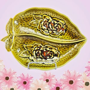 以下が含まれることがあります： 金色がかった黄色の、葉の形をした2つのセクションに分かれたサービングディッシュ。赤とオレンジ色の中心を持つエンボス加工のバラが特徴で、銀色の金属製のスタンドで支えられています。この皿はおそらくセラミック製です。