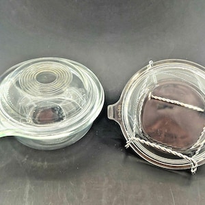 May include: Two clear glass Pyrex casserole dishes with lids. One dish has a handle and a lid with concentric circle design. The other dish has a metal handle and a dark brown interior.